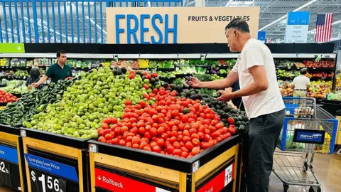 AFP/Getty A man in a white T-shirt holds an avocado while standing in the produce section before a pile of tomatoes, avocadoes and other vegetables at a Walmart in Rosemead, California, on April 11, 2025.