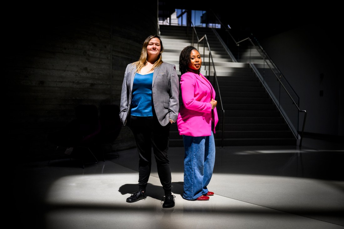 Nita Akoh wearing a pink blazer and blue pants and Jay Nakhai wearing a grey blazer and black pants standing back to back under a spotlight-type light in a darkened room.