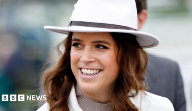 Princess Eugenie smiles off to the side of the camera while walking along. She has wavy brown hair and blue eyes. She is wearing a white coat and white hat.