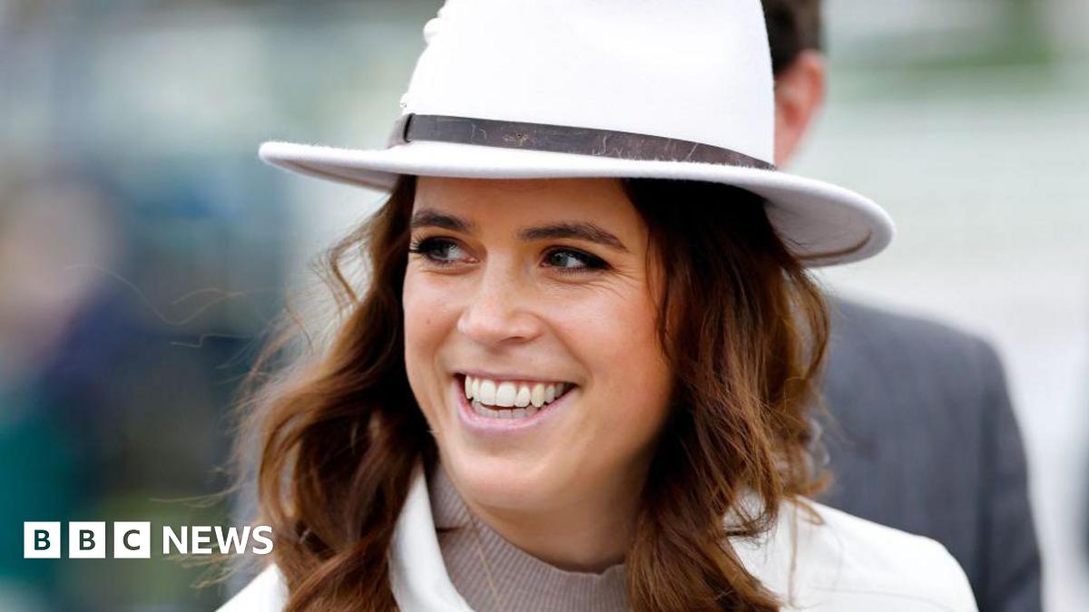 Princess Eugenie smiles off to the side of the camera while walking along. She has wavy brown hair and blue eyes. She is wearing a white coat and white hat.