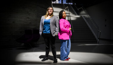 Nita Akoh wearing a pink blazer and blue pants and Jay Nakhai wearing a grey blazer and black pants standing back to back under a spotlight-type light in a darkened room.