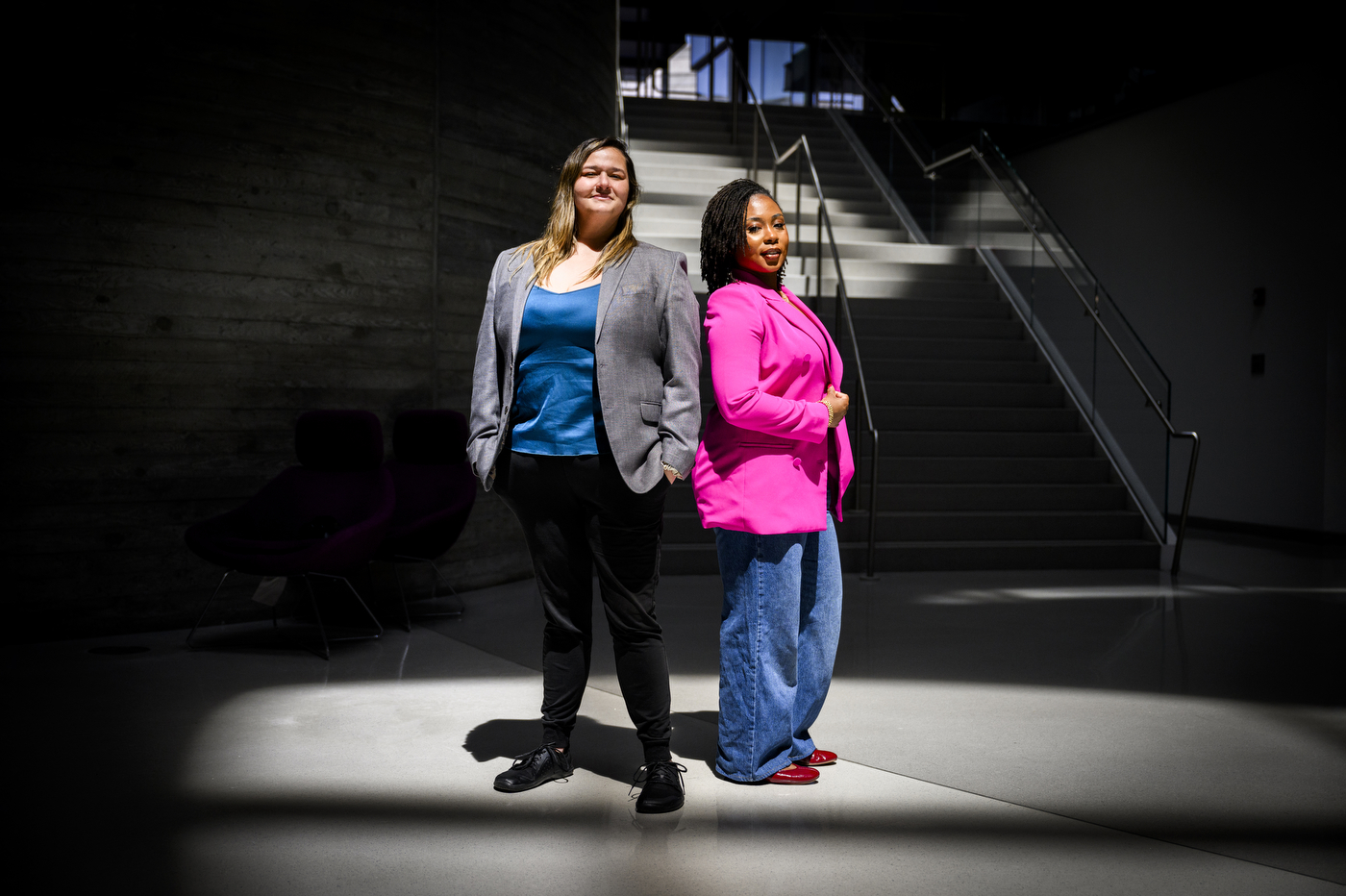 Nita Akoh wearing a pink blazer and blue pants and Jay Nakhai wearing a grey blazer and black pants standing back to back under a spotlight-type light in a darkened room.