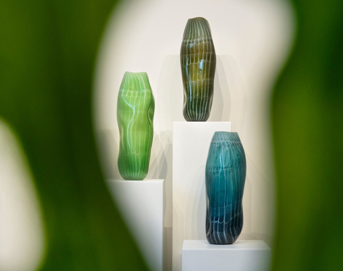Trois vases en verre de différentes teintes de vert et bleu, disposés sur des socles blancs, vus à travers un feuillage flou.