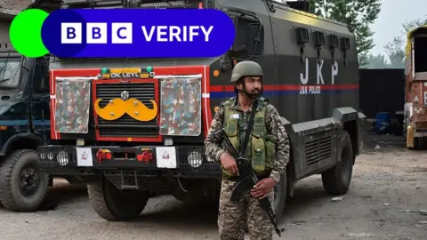 Getty Images/SOPA Images An Indian soldier stands guard. He is armed with a rifle and is wearing army fatigues. In the background is a large truck.