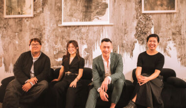 A photograph of four people sitting on a couch in front of a wall of artwork.