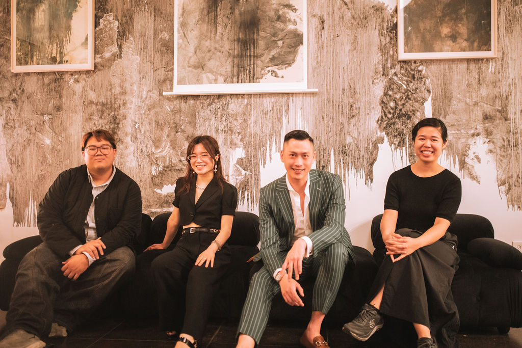 A photograph of four people sitting on a couch in front of a wall of artwork.