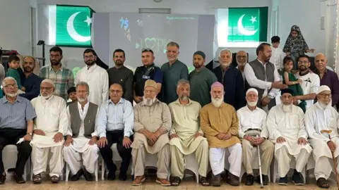Atiff Ghfar A group of people from the Swindon Pakistan Association pose for a photo. There is a row of people sititng on chairs and another row of people standing behind them. There are two Pakistani flags in the windows on the wall.