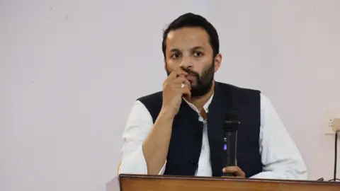 Instagram/Ali Khan Mahmudabad The picture shows Ali Khan Mahmudabad wearing a white shirt with a black sleeveless jacket standing besides a podium with a mic in his hand