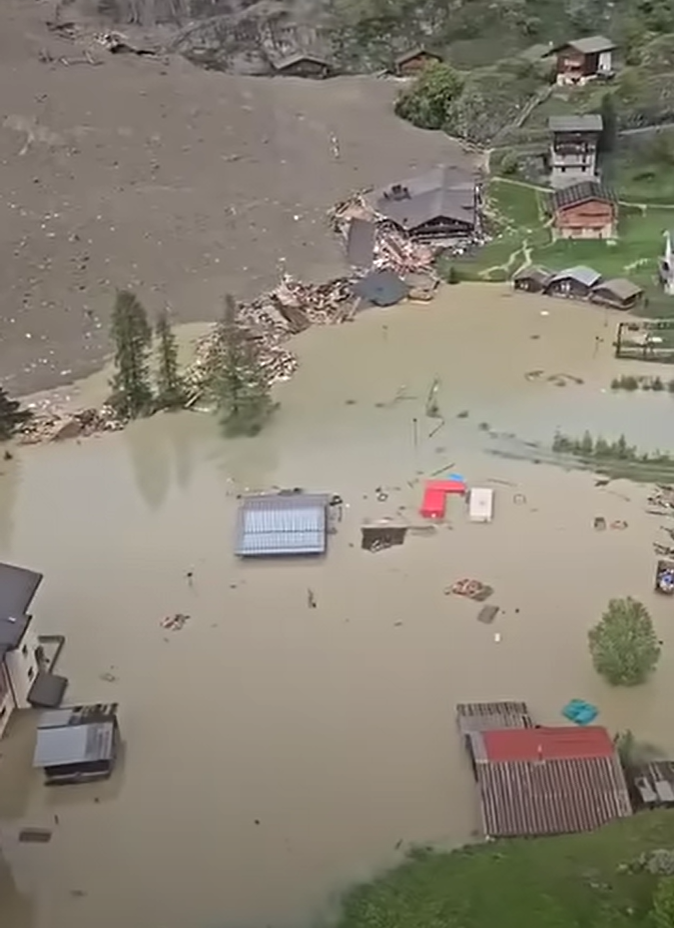Überflutete Landschaft mit beschädigten Häusern und Trümmern im Wasser, umgeben von Erdrutschspuren.