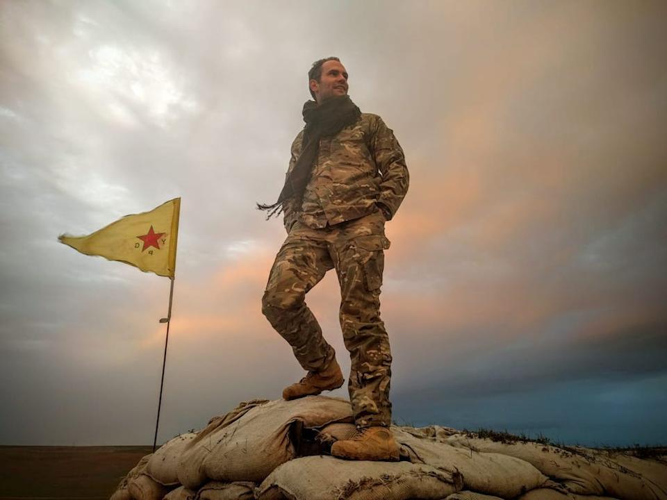 Macer Gifford/Harry Rowe in a military uniform, standing on a pile of sandbags against a rosy clouded sky, with the yellow flag of the People's Defense Units of Syria beside him.