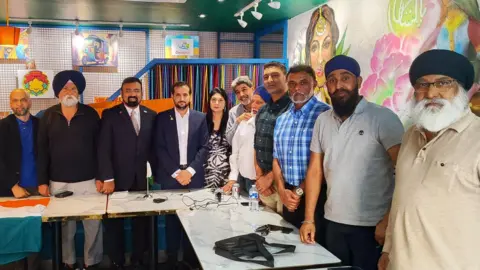 Rana Basharat Ali Khan A group of people, largely men, stand around a table in what appears to be a meeting room. Most are looking at the camera with a serious expression. 