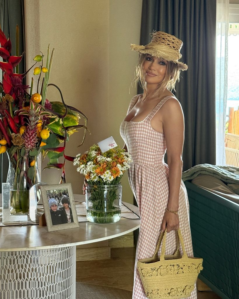 In this warmly lit indoor photograph, Jennifer Lopez stands gracefully beside a round table adorned with vibrant floral arrangements and a framed photograph of two children. She wears a soft pink and white gingham milkmaid-style dress that hugs her figure and flares gently at the bottom. Her hair is styled in relaxed waves and partially tucked under a wide-brimmed raffia hat. On her arm, she carries a straw-like raffia tote bag with intricate floral crochet detailing. The ambiance is breezy and summery, with natural light streaming in from a window in the background, highlighting the seaside setting beyond the room. Her expression is serene, radiating a casual elegance.