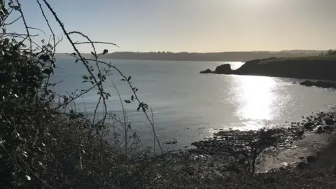 BBC A cove is seen with the sun shimmering on the sea and a rock formation in the background