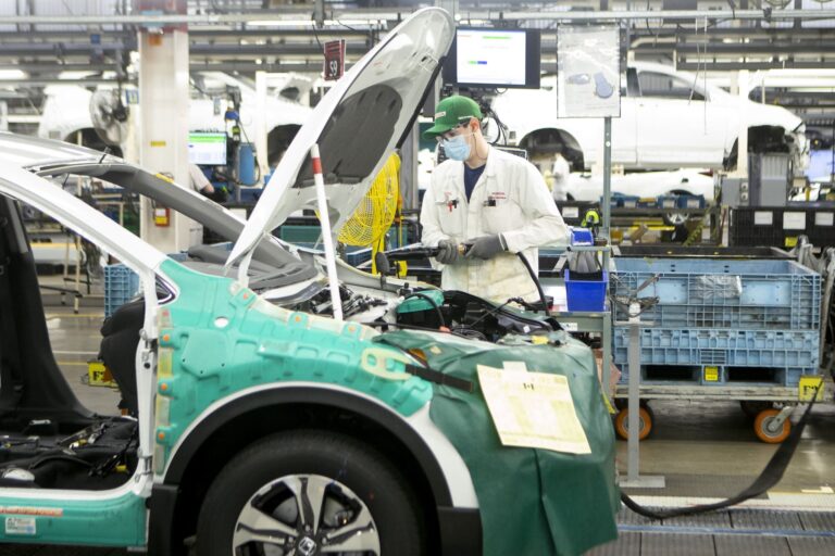 An autoworker in a white overcoat, green hat, face mask and protective glasses works on a partially constructed vehicle on a Honda assembly line.
