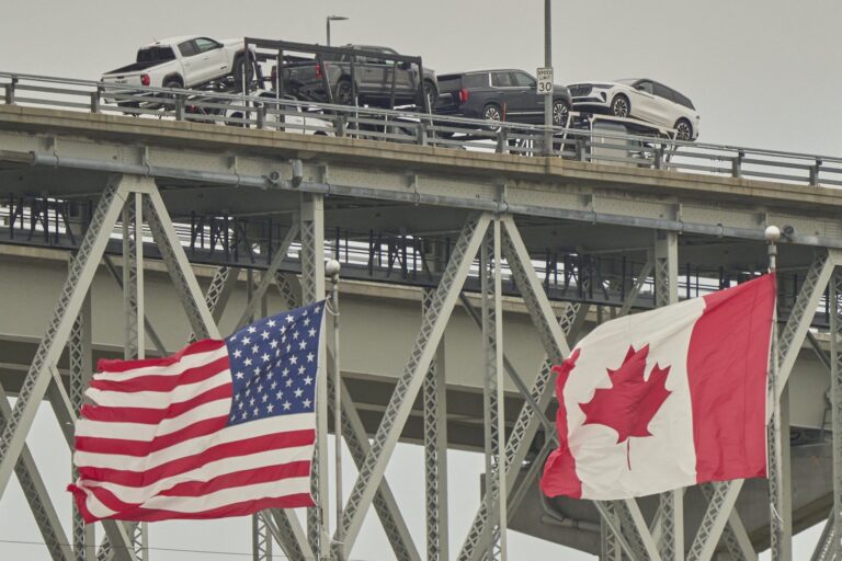 A low-angle shot of a truck carrying vehicles across the bridge at the Canada-U.S. border in Sarnia, Ont. The U.S. and Canadian flags are flying in the foreground.