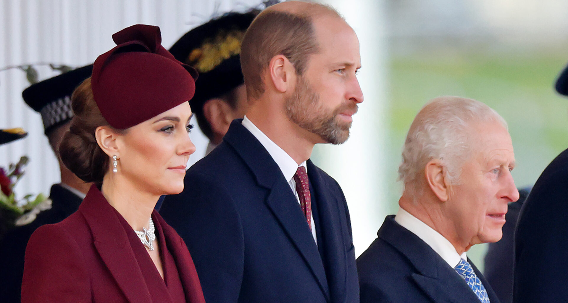 Kate Middleton wears a burgundy coat and matching hat, Prince William wears a navy coat and red tie, and King Charles wears a navy coat with a white shirt and blue tie