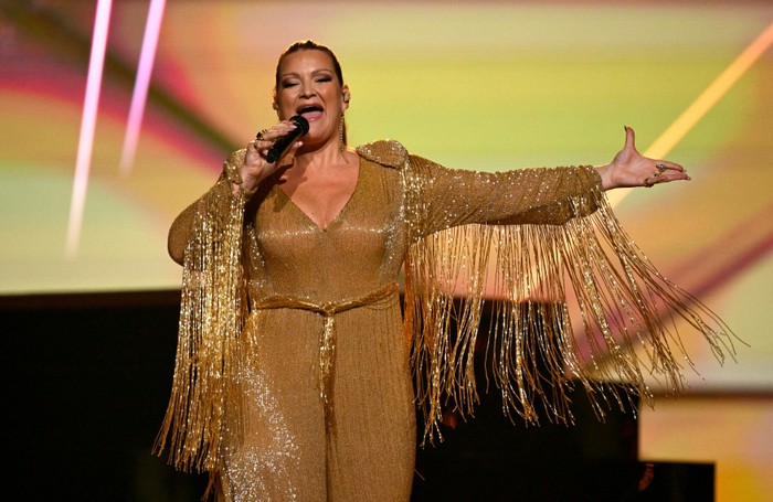 Hera Björk wears a gold sparkly fringed jumpsuit on stage and sings into her microphone with her other arm out stretched to the side.