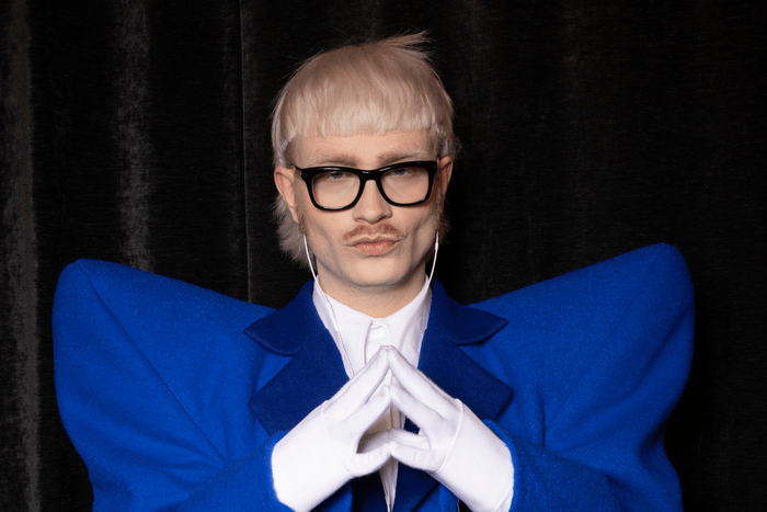Joost Klein poses at a press meeting before Eurovison The Concert. He is wearing a dark blue suit with shoulder pads and has bleached blonde hair.