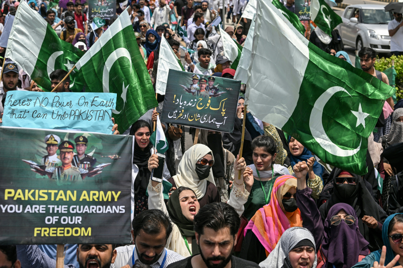 A crowd of dozens of protesters waves signs and green-and-white Pakistani flags at a demonstration.