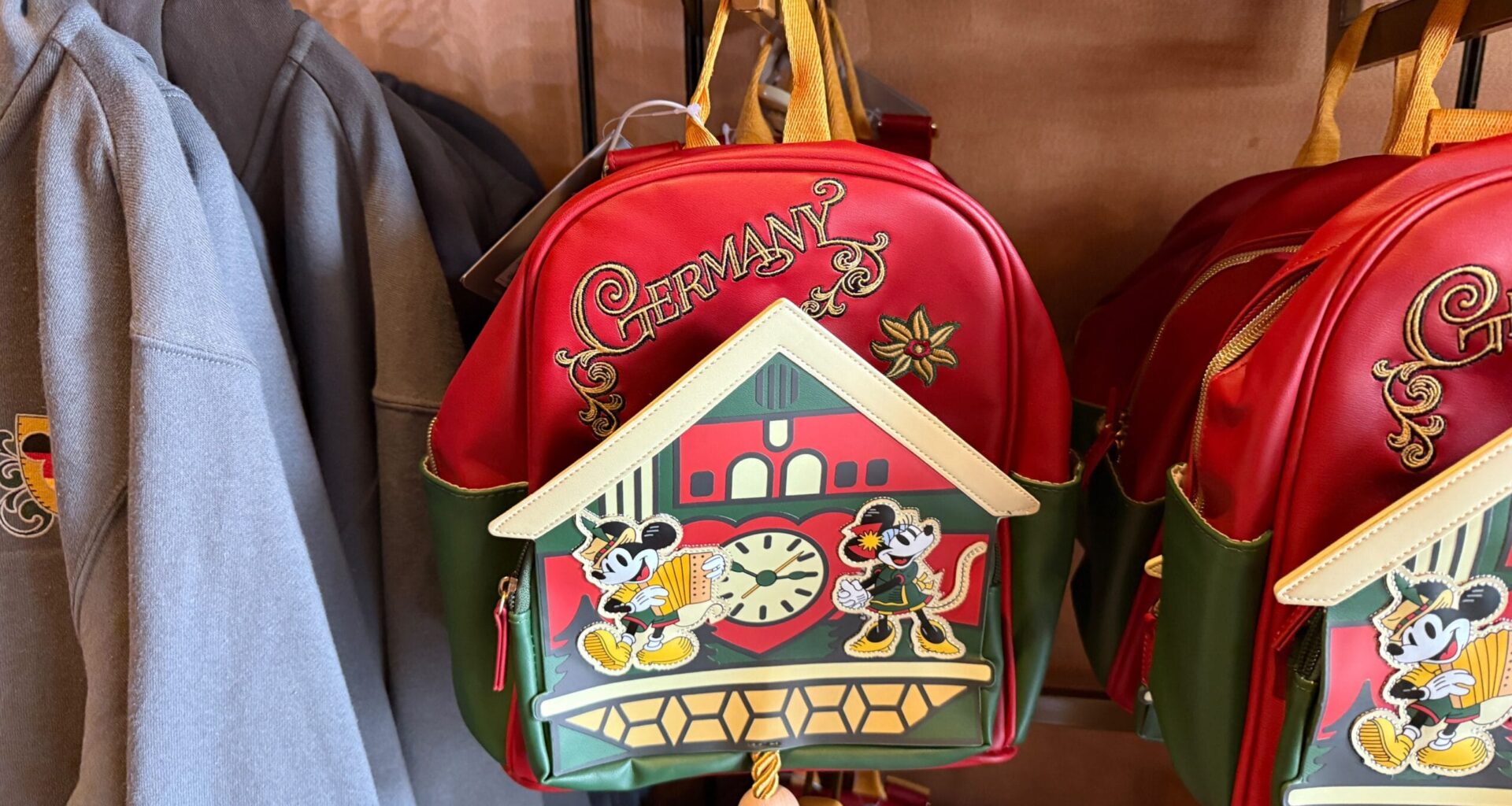 Red and green backpack with "Germany" text and cartoon mice by a cuckoo clock, hanging on a store display.