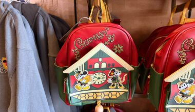 Red and green backpack with "Germany" text and cartoon mice by a cuckoo clock, hanging on a store display.