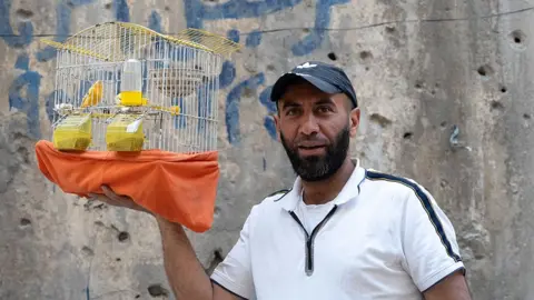 Rolf Andreason/BBC Yasir stands outside a bullet-marked building holding a cage containing his pet bird 