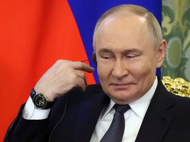 Vladimir Putin, con gesto sonriente, se lleva la mano derecha a la oreja derecha, en la que tiene un audifono. 