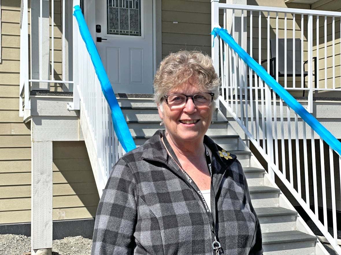 A woman with short grey hair and a plaid shirt stands in front of a newly built tan house.