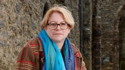 Neil Spence Prof Marion Gibson, sitting outside a historic stone building. She has fair, centre-parted hair and is wearing tortoiseshell glasses and a turquoise scarf over a thick red, black and brown checked coat.