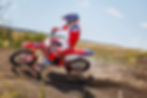 Motorcyclist in red and blue gear rides a dirt bike on a trail, kicking up dust under a clear sky. Fields and flowers in the background.