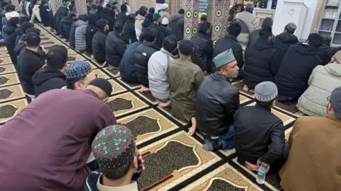 Abdul Malik A large group of men kneel on prayer mats in a mosque. Some are wearing a kufi or taqiyah, and many are wearing winter coats. They are all facing away from the camera, and appear to be in conversation with each other. 