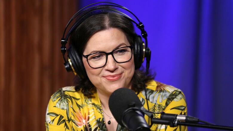 A smiling woman wearing glasses and over-ear headphones sits in front of a studio microphone.