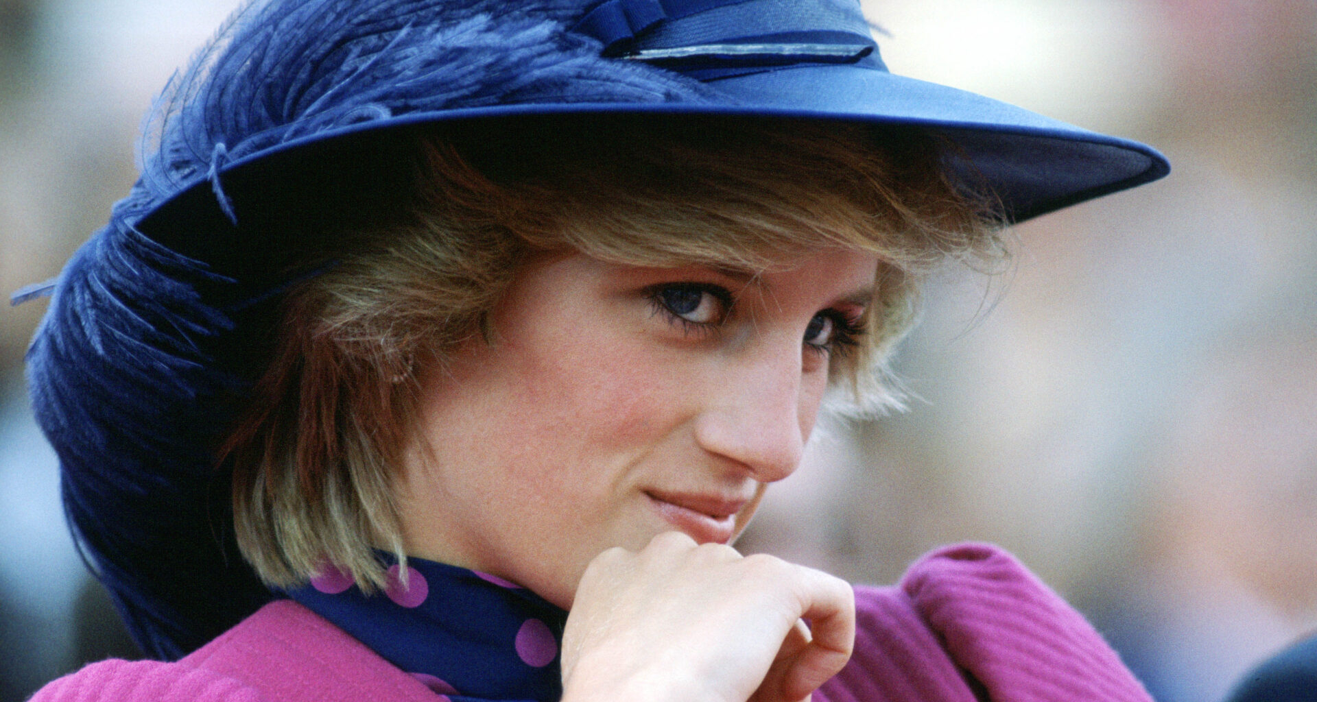 Princess Diana wearing a blue feathered hat and pnik dress leaning her chin on her hand