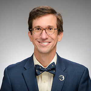 Man with brown hair and glasses, wearing a navy suit with cream colored shirt and navy patterned bow tie, smiles against a gray background.