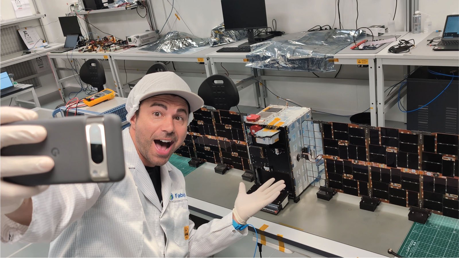 A smiling person in a lab coat and gloves takes a selfie in a laboratory, gesturing toward a small satellite or spacecraft model on a workbench with equipment and computers in the background.