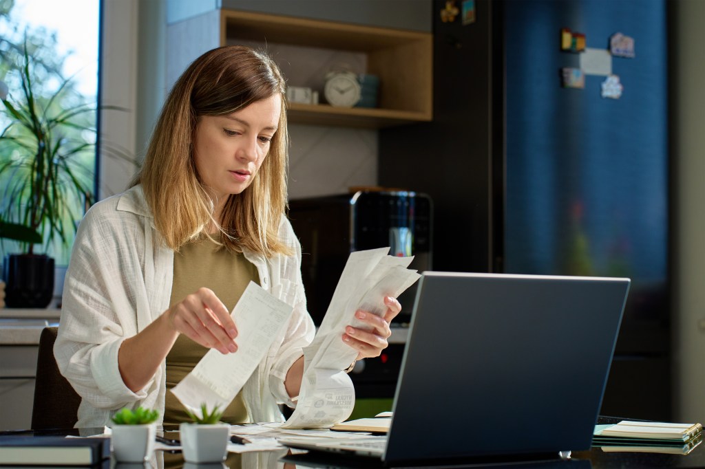 Woman stressing over bills, seated at kitchen table with laptop and paper documents, managing personal finances