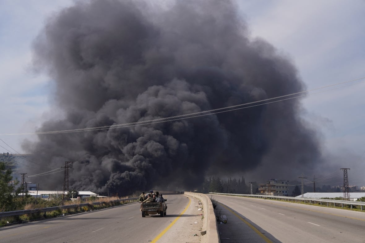 A military vehicle travels along a deserted highway, toward a large plume of black smoke.