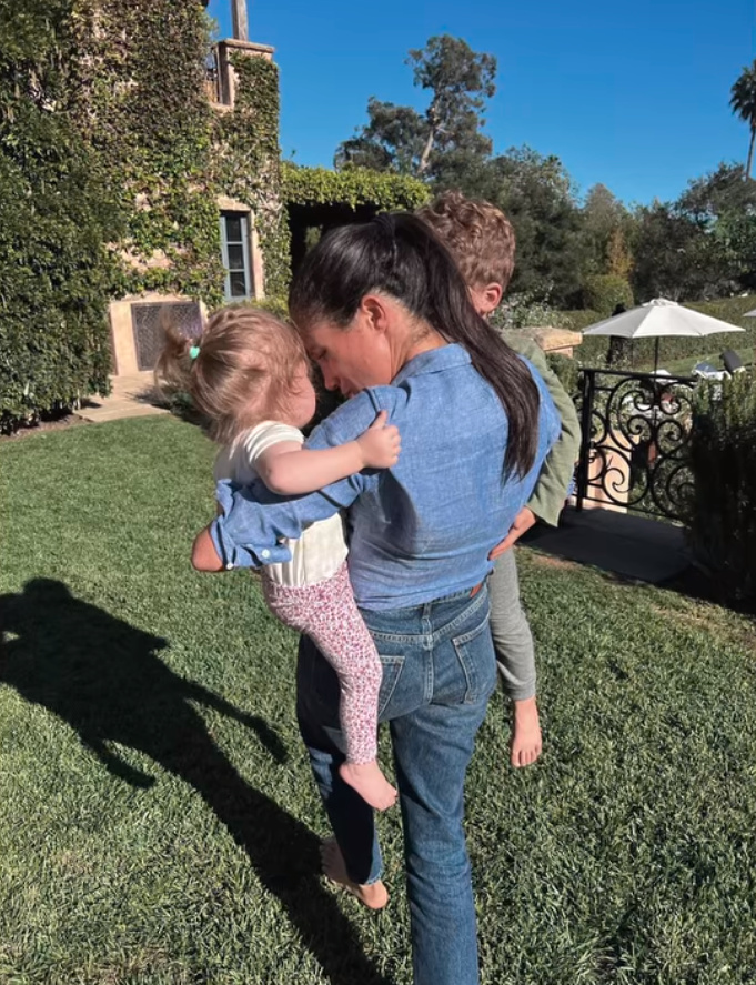 Meghan Markle carrying her two children on a lawn.