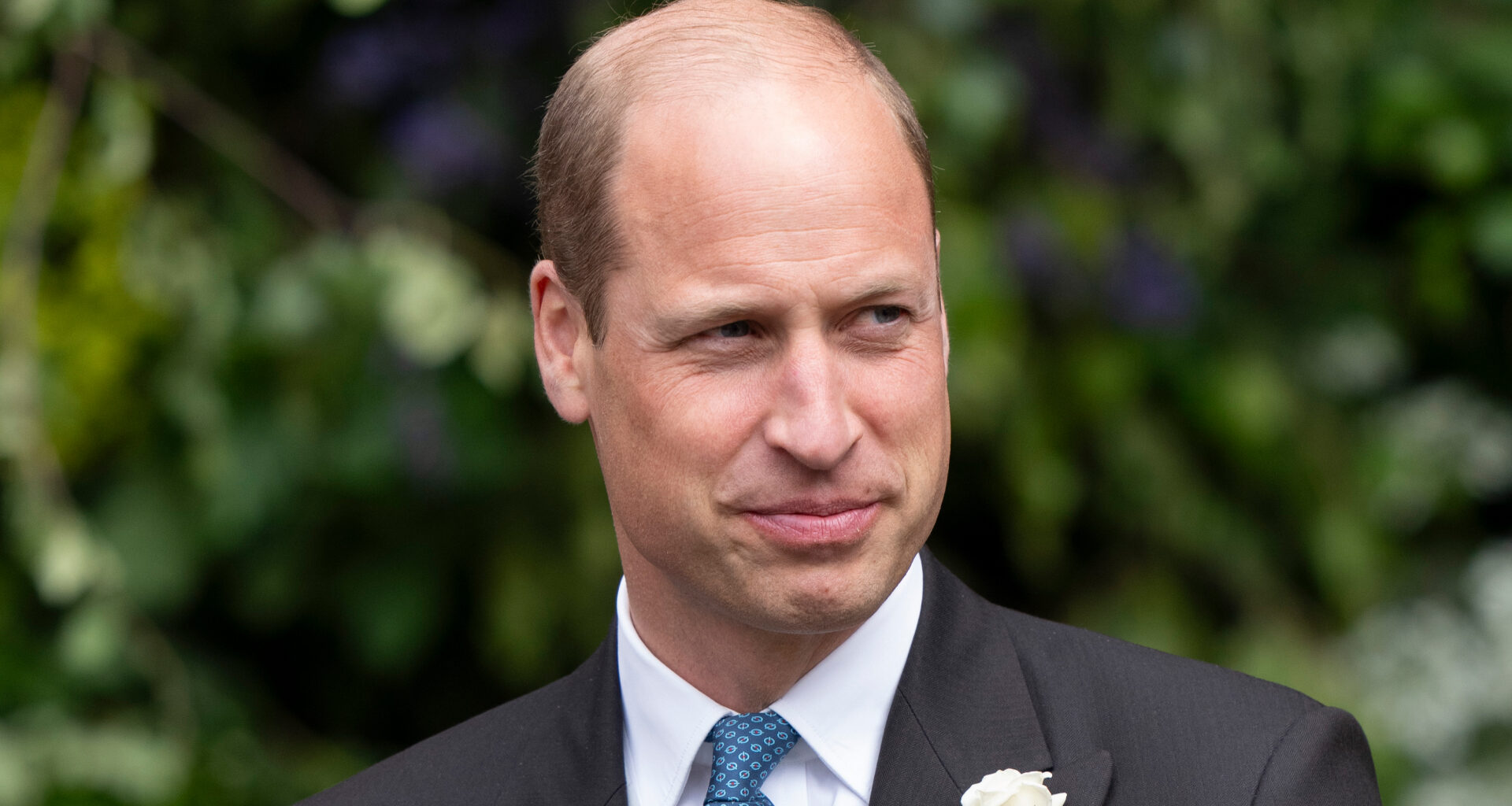 Prince William wearing a suit with a white flower and smiling in front of trees at the Duke of Westminster's 2024 wedding