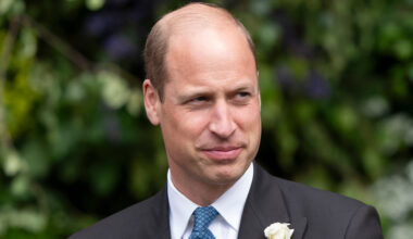 Prince William wearing a suit with a white flower and smiling in front of trees at the Duke of Westminster's 2024 wedding