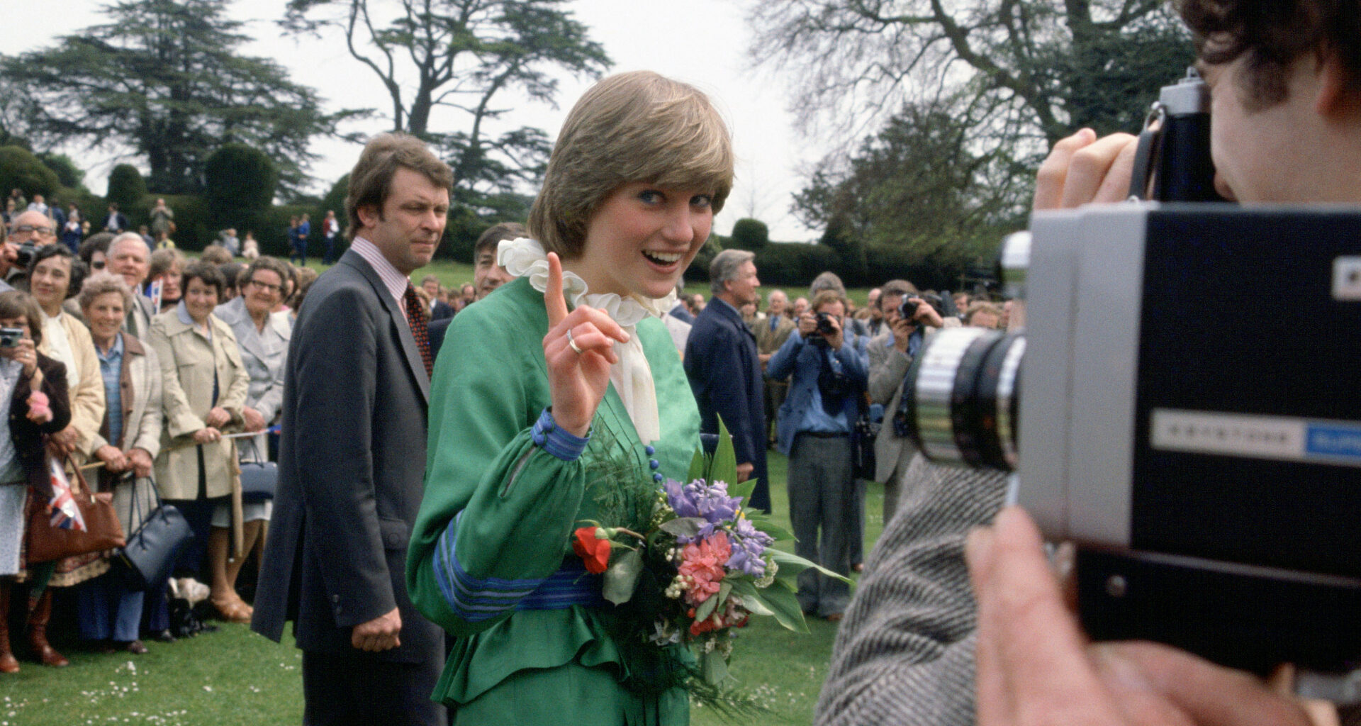 Princess Diana wearing a green dress waving her finger at the camera as a man takes a video of her