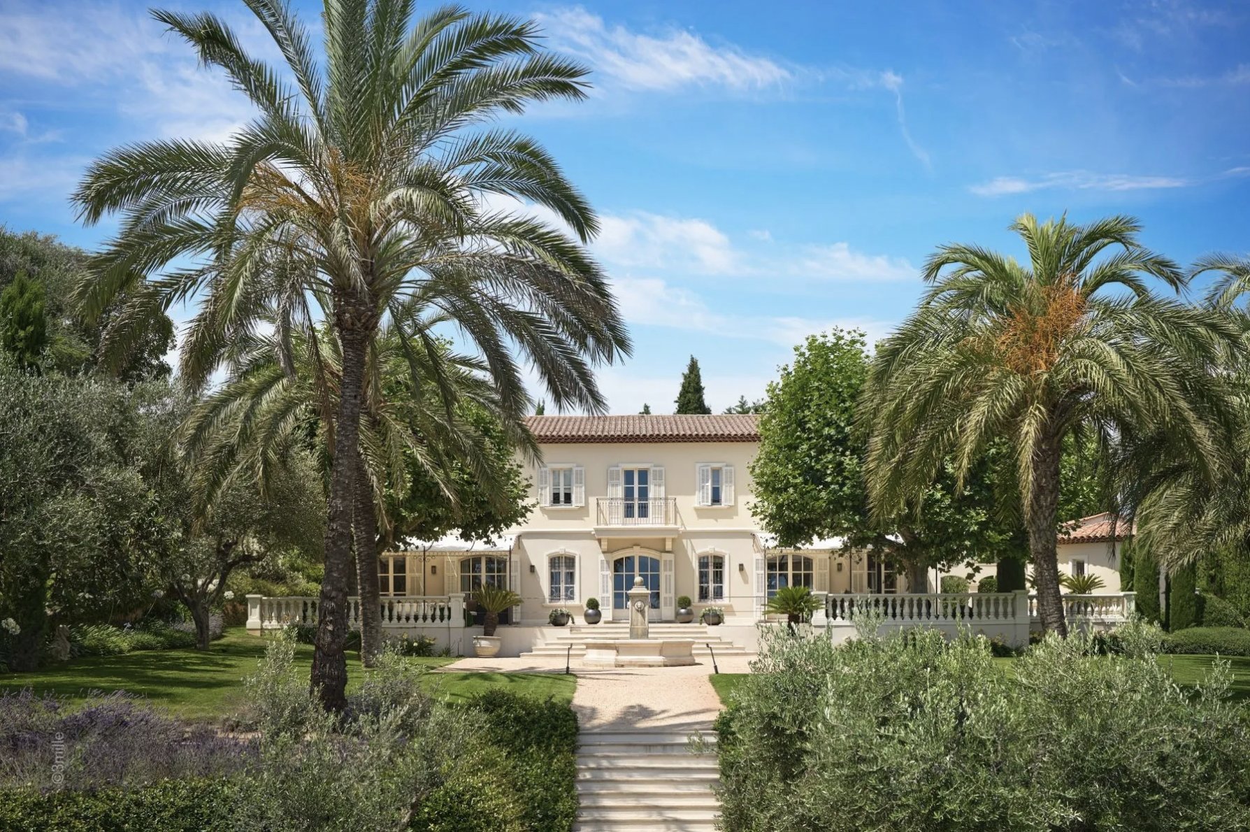 Large, light beige house with a fountain in the front, surrounded by palm trees and landscaping.