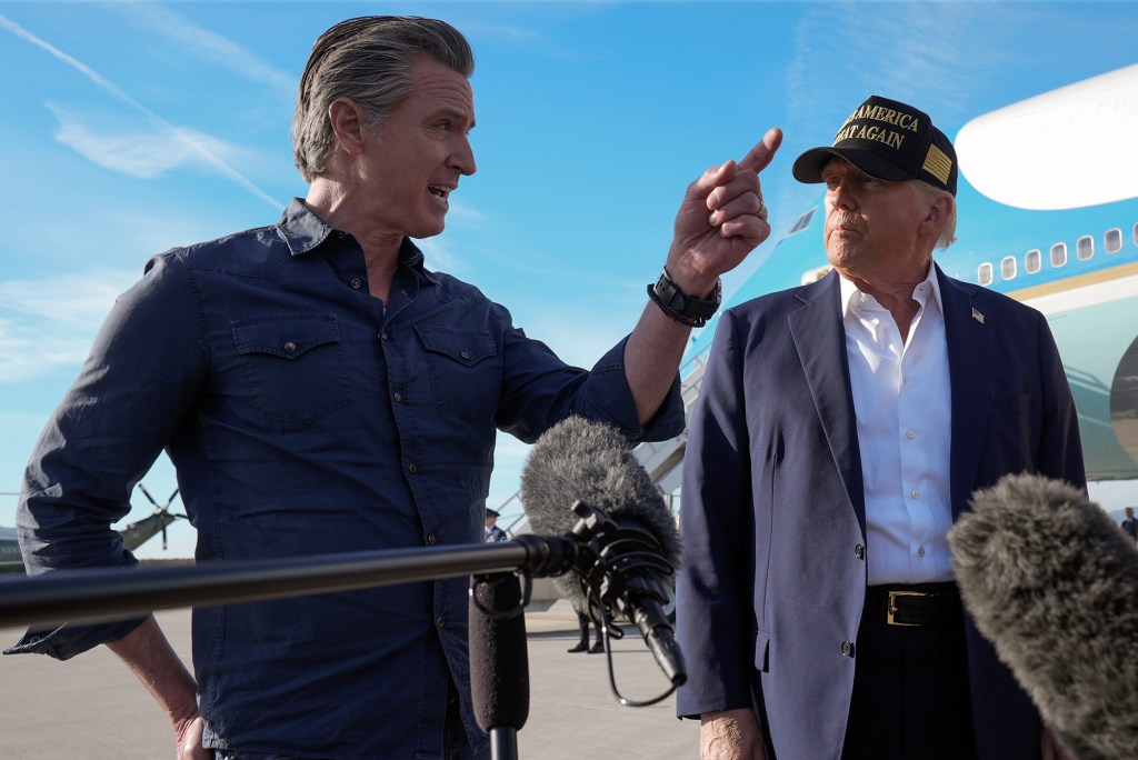 Two individuals stand on an airport tarmac, engaged in an intense discussion. One person, wearing a dark button-up shirt, gestures emphatically while speaking. The other individual, dressed in a blazer and a cap with gold lettering, looks on attentively. A large aircraft and a clear blue sky serve as the backdrop, with microphones visible in the foreground.