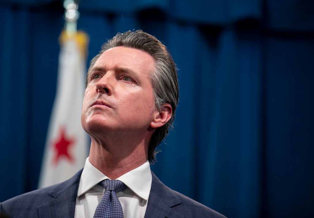 California Governor Gavin Newsom looks upward with a serious expression during a press conference. He is dressed in a suit and tie, standing in front of a blue curtain with a partially visible California state flag in the background.
