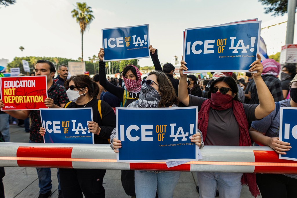 A group of protesters stands behind a red-and-white barricade holding signs that read “ICE out of LA!” and “Education not deportation.” Many participants wear face coverings, sunglasses, and masks. The protest appears to be taking place outdoors in a city setting with palm trees and buildings in the background.