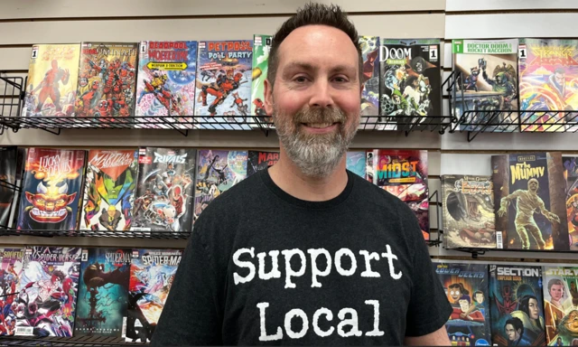 A man smiling at the camera wearing a black tshirt that says support local. He stands in front of shelves full of comic books