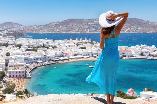 A woman in a blue dress stands on a cliff overlooking Mykonos