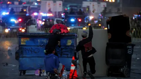 Reuters Two men crouch behind large bins with riot police in the distance