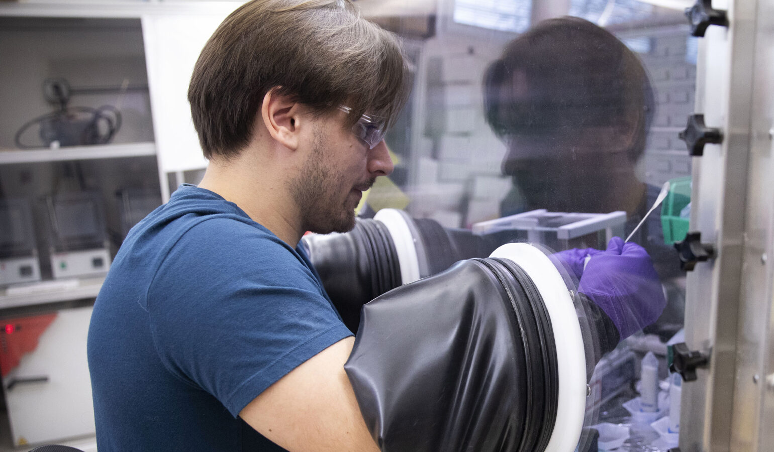 A person uses a glove box to handle materials in a laboratory, wearing safety glasses and a blue shirt.