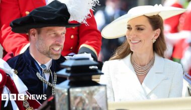 The Prince and Princess of Wales in a horse carriage. The prince wears ceremonial robes and hat, the princess is in a white jacket and white hat with a pearl necklace.